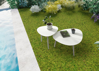Rowa small outdoor side table with rounded pebble-inspired shape, featuring a plant and coffee cup, on green grass near pool.